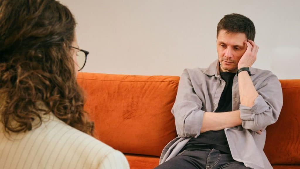 A man sitting on an orange sofa, holding his head while talking to a woman.