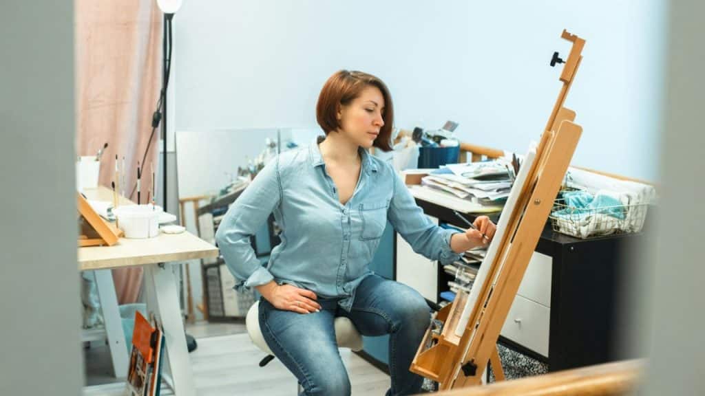 Woman in denim shirt and jeans painting on an easel in a cluttered studio.