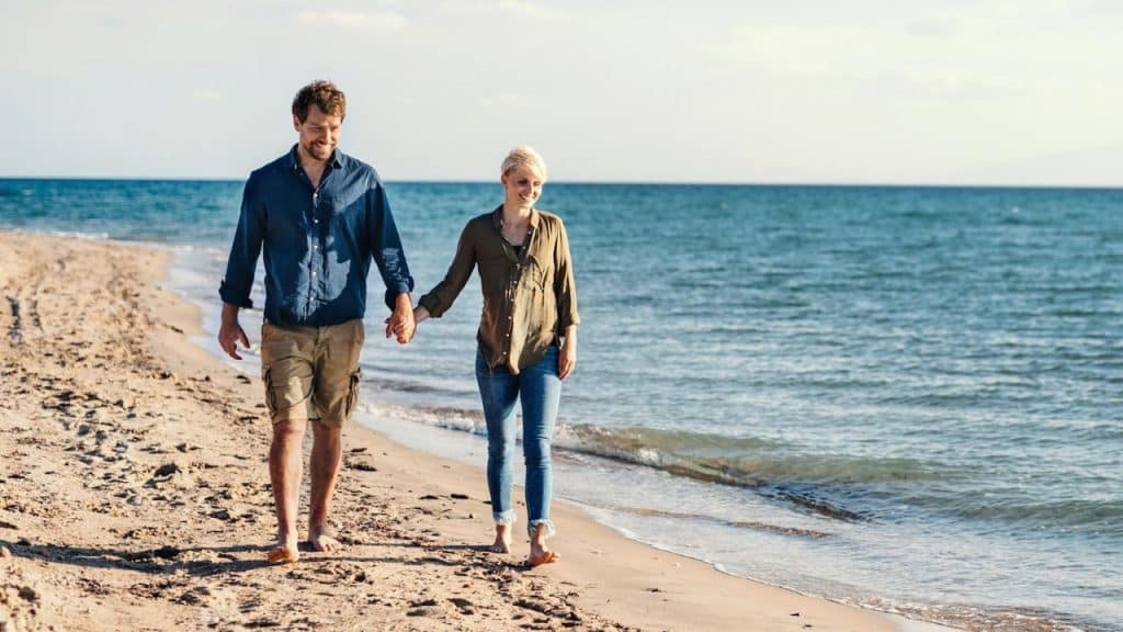 A smiling couple walks hand-in-hand along a sandy beach near the ocean.