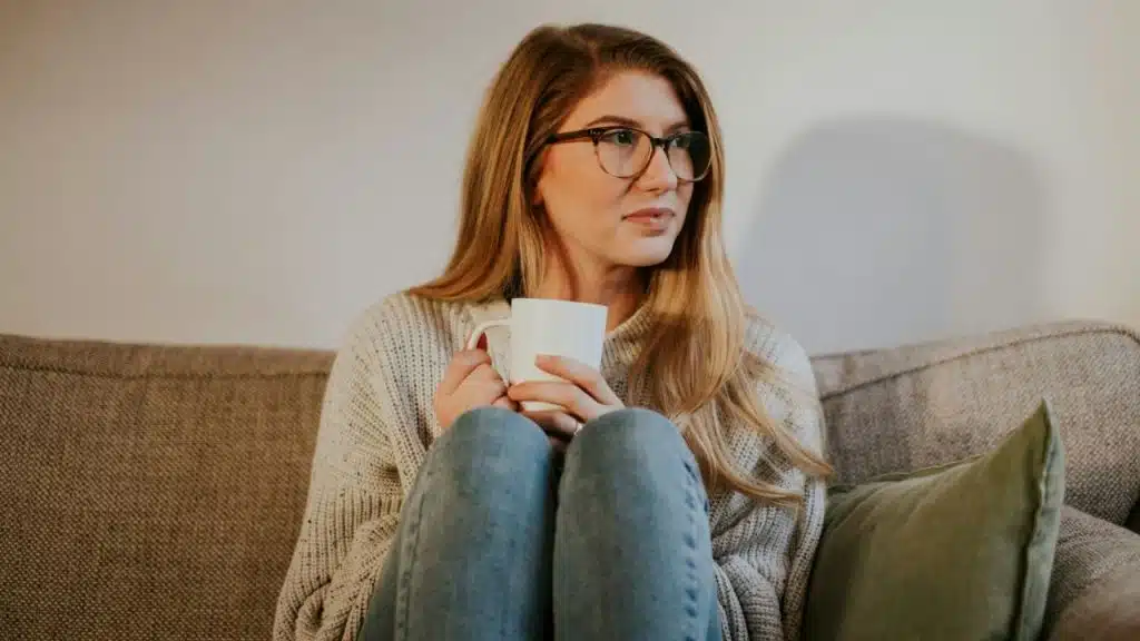 A woman wearing glasses and a sweater sits on a couch holding a white mug.