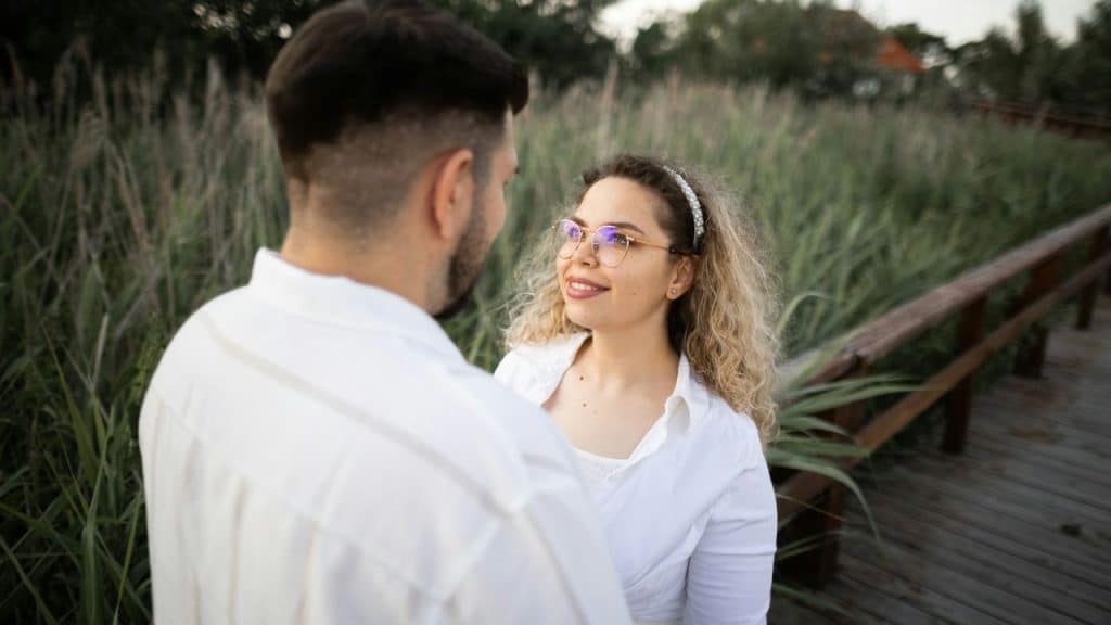 Smiling woman with glasses looks up at a man on a wooden path surrounded by tall grass.