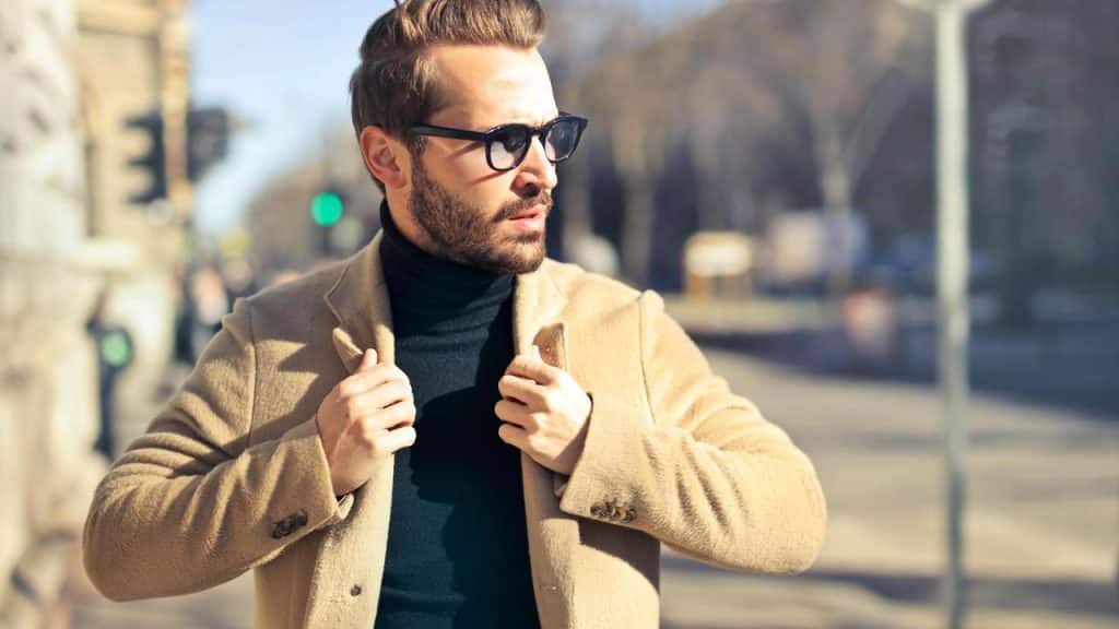 A man in sunglasses adjusting his beige coat while standing outdoors.