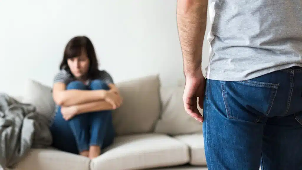 Man's back and jeans are in sharp focus in the foreground, while a woman sits in the background on a couch, hugging her knees and looking upset.