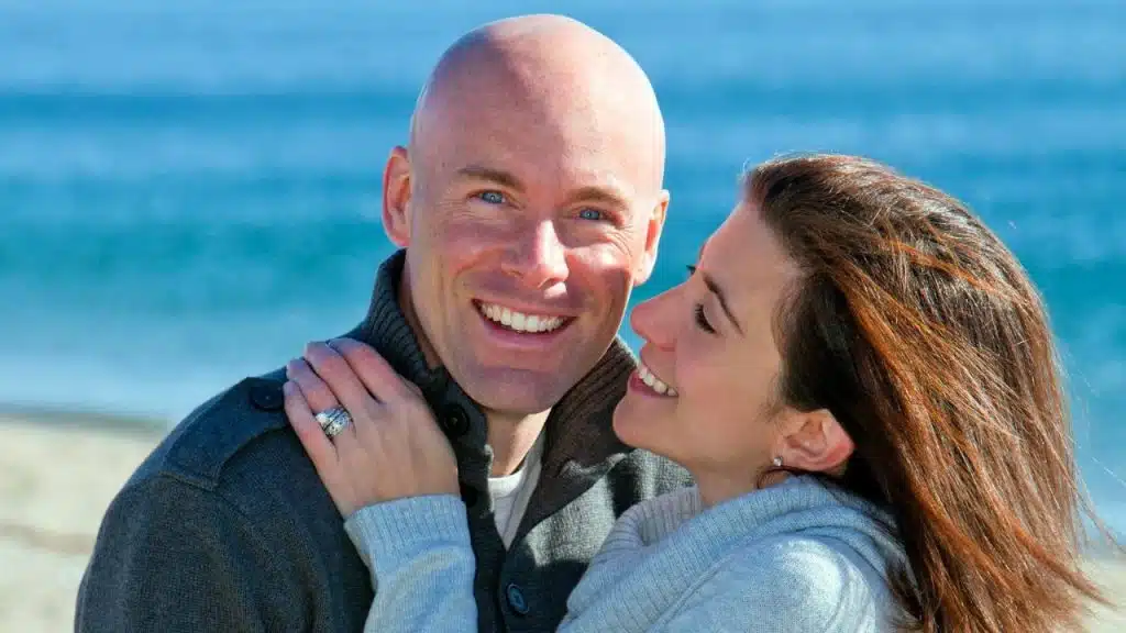 Smiling bald man and woman embracing on a sunny beach near the ocean.