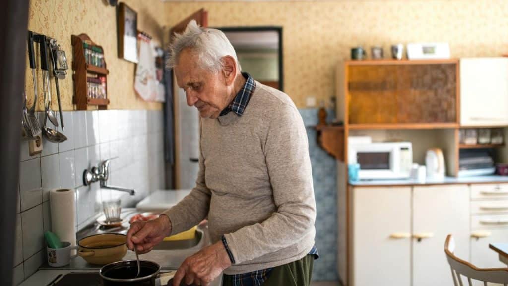 Elderly man stirring a pot on a stovetop in an old-fashioned kitchen.