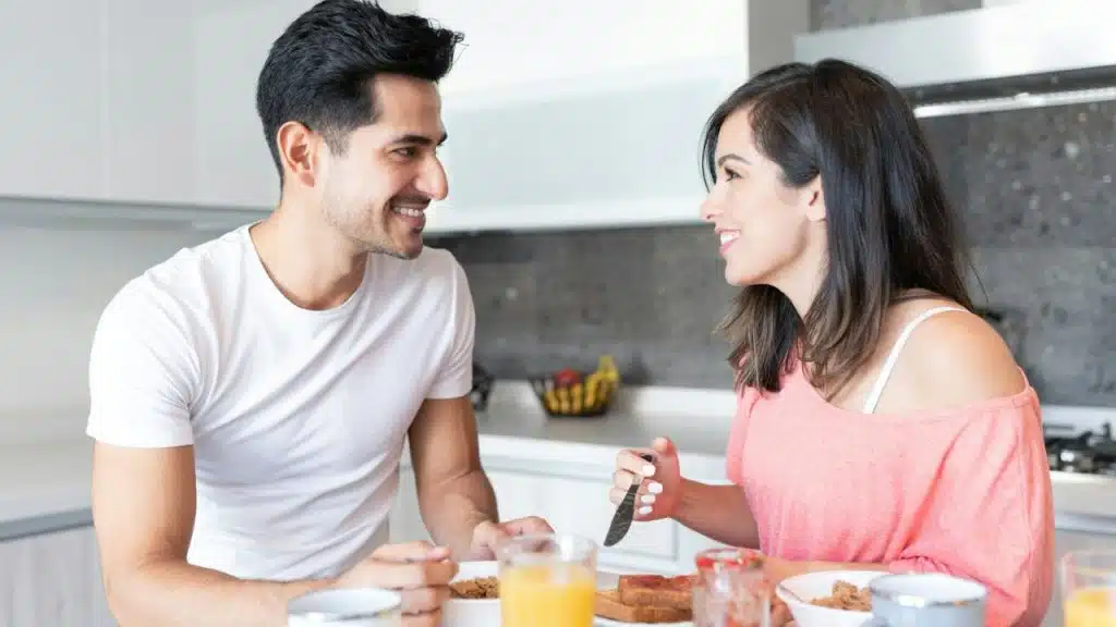 Man and woman smiling at each other across a table while eating breakfast in a kitchen.