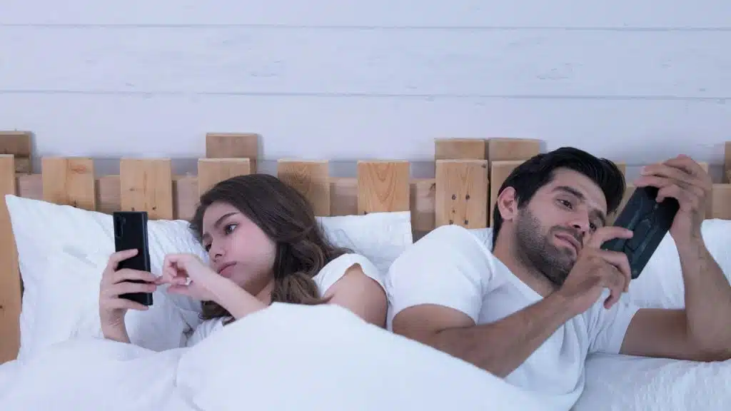A couple lying back-to-back in bed, each absorbed in their smartphone.