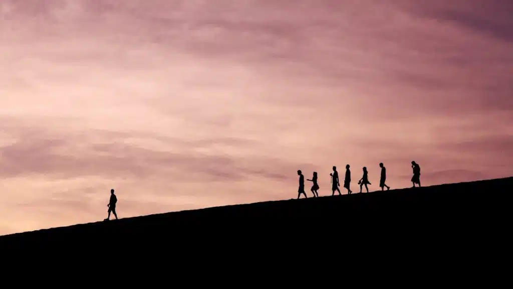 Silhouette of a single person walking up a hill in a vast, open landscape with a colorful sunset sky, away from a line of several other people also walking.