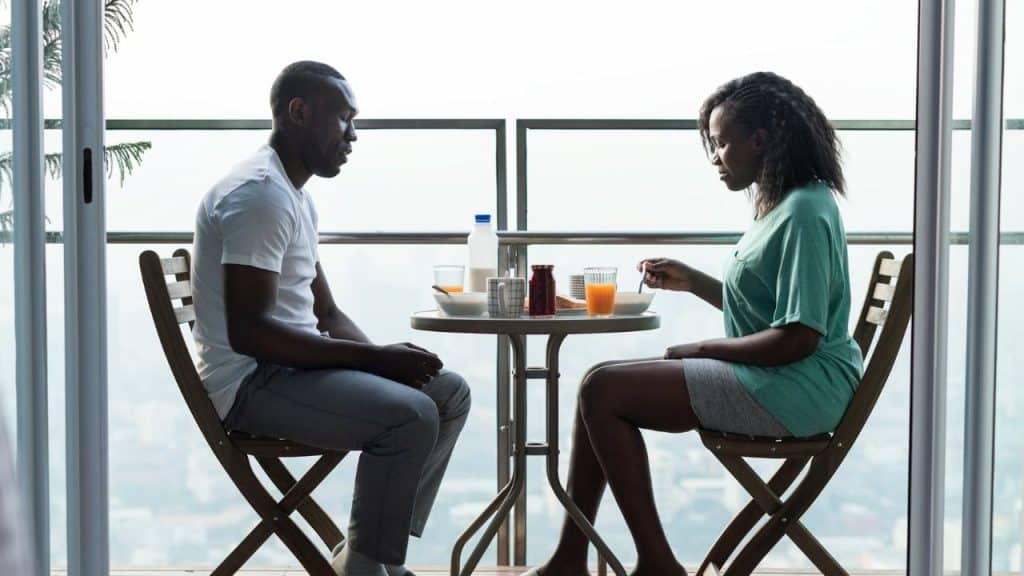 A man and a woman sit facing each other at a small table on a high-rise balcony.