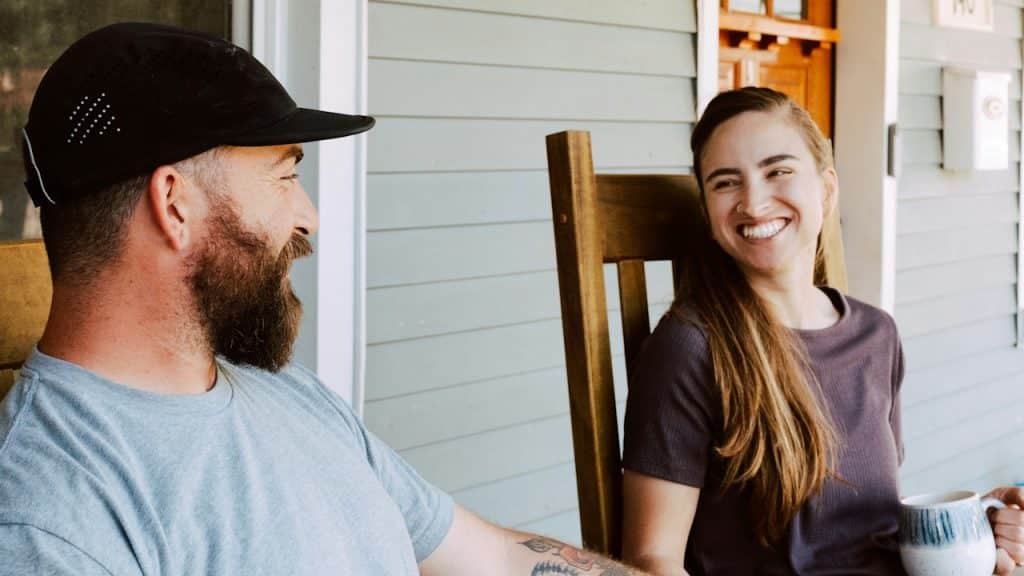A man and a woman sit in rocking chairs on a porch, sharing a laugh while the woman holds a mug.