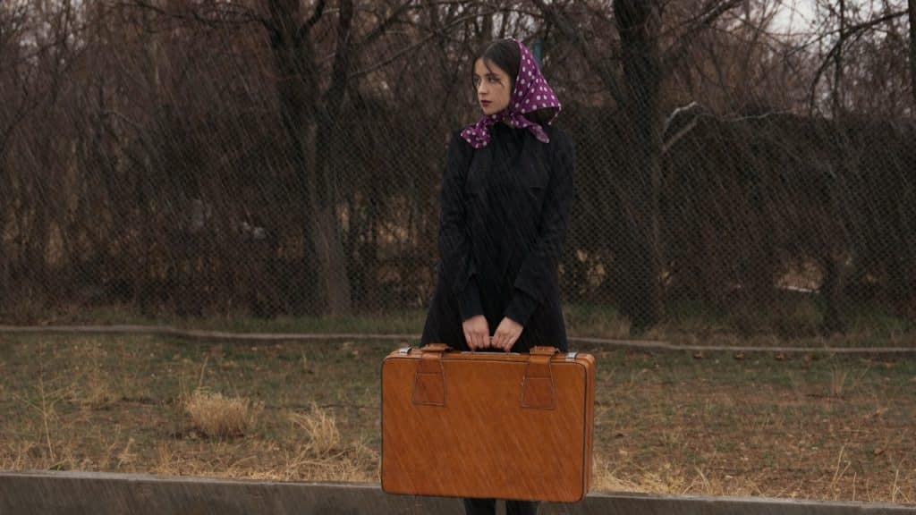 A woman standing on a road in rain with a piece of luggage.