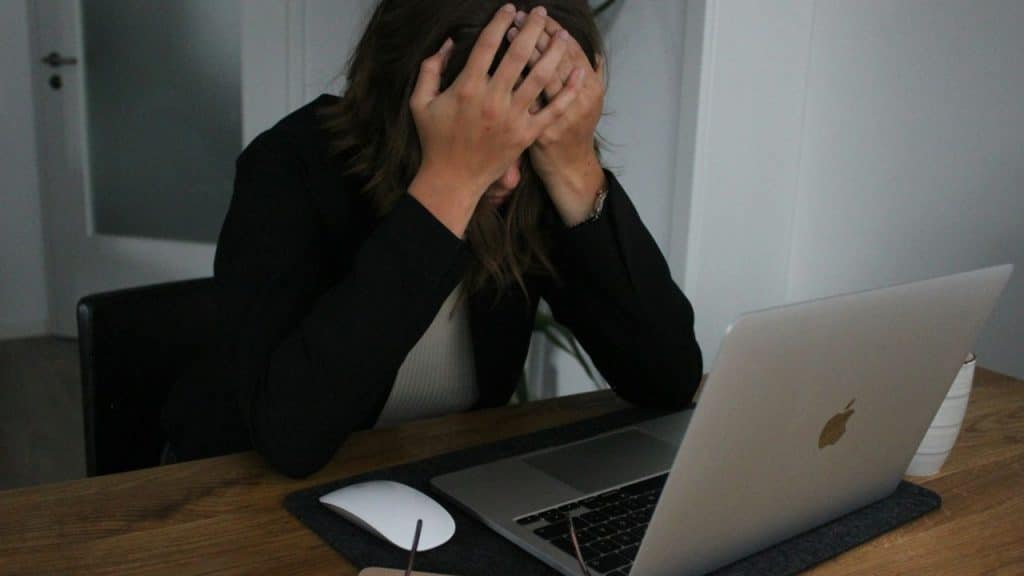 Woman in black blazer with hands on her head looks down at a silver laptop.