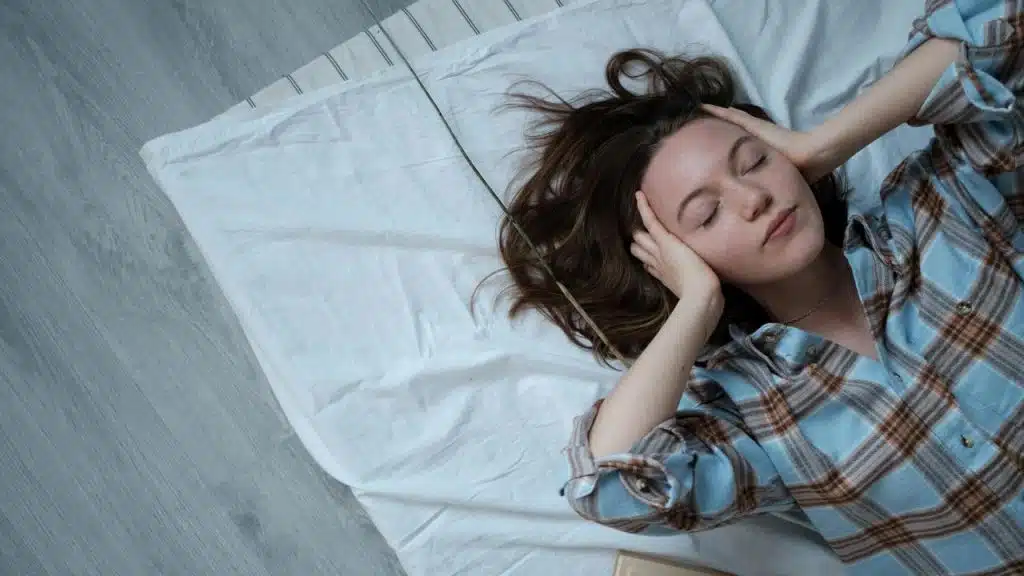 A woman lying on a bed with her hands on her head and eyes closed.