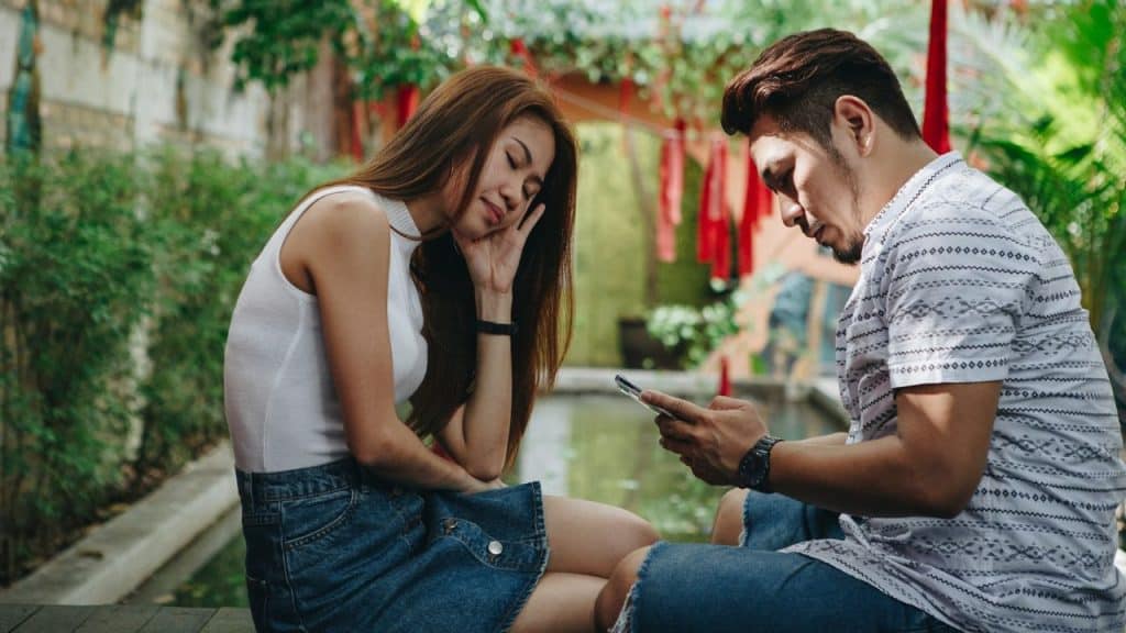 A man stares intently at his phone while sitting next to a woman leaning her head on her hand.