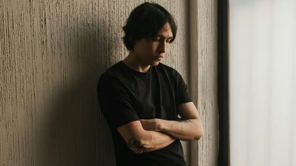 Young man in black shirt leans against a textured wall with arms crossed.