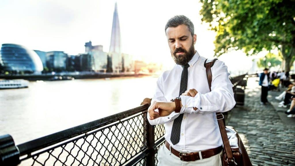 Man with a beard and tie checking his watch by a river in a city.