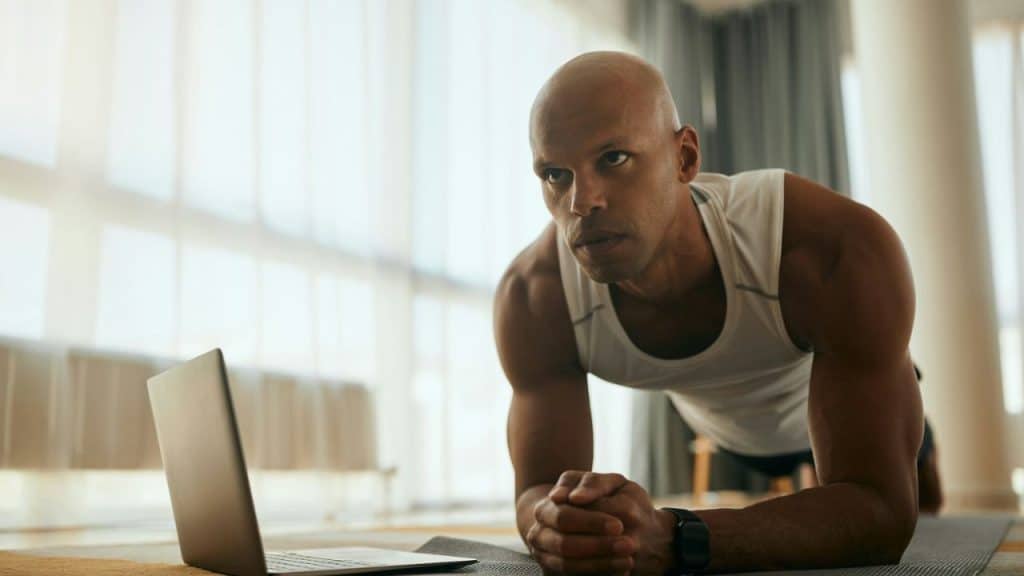 Bald man in a white tank top doing a plank exercise near a laptop.