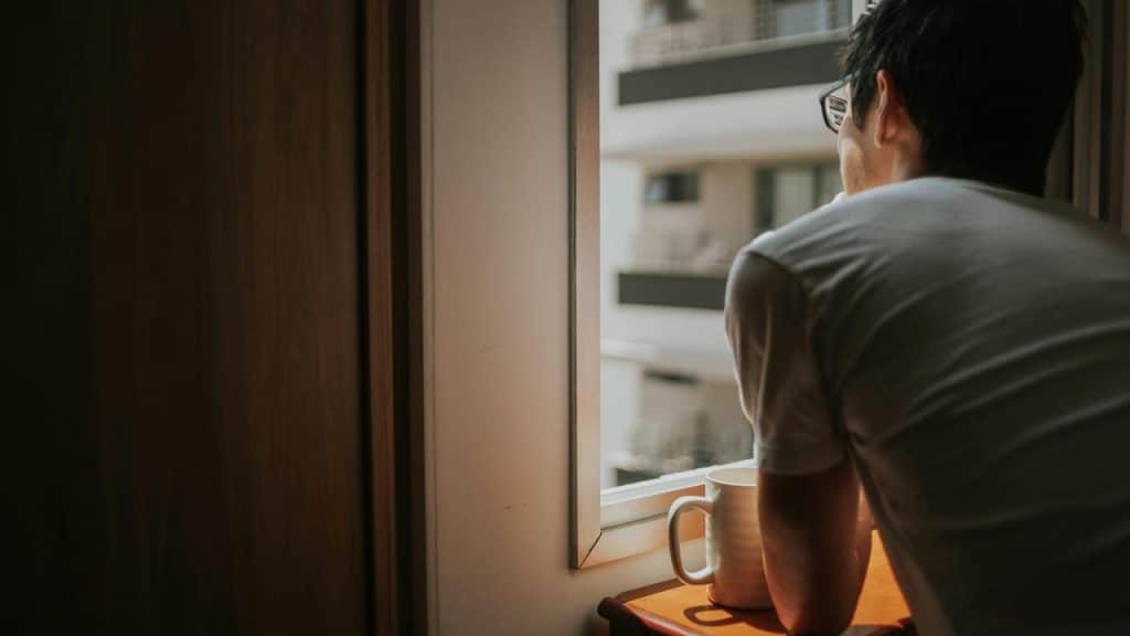 A person standing by a window holding a mug and looking outside at nearby buildings.