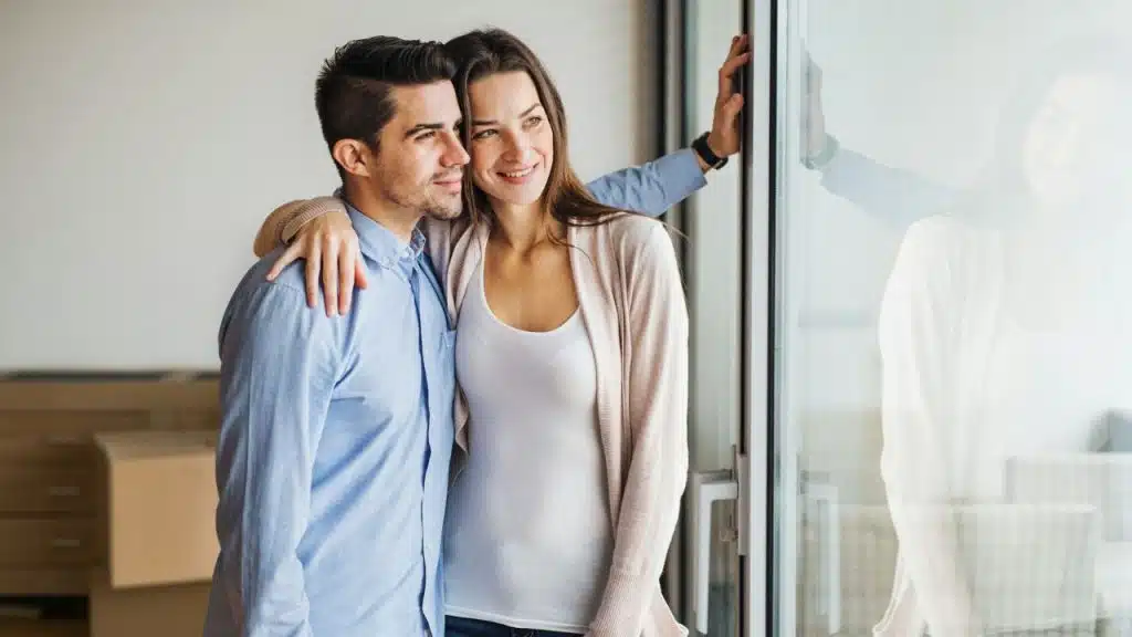 A man and woman stand by a window together, smiling and embracing while looking outside.