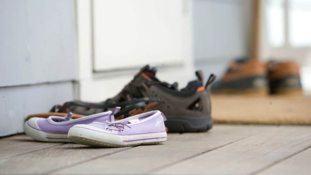 Several pairs of shoes, including purple sneakers, sit on a wooden porch near a door.