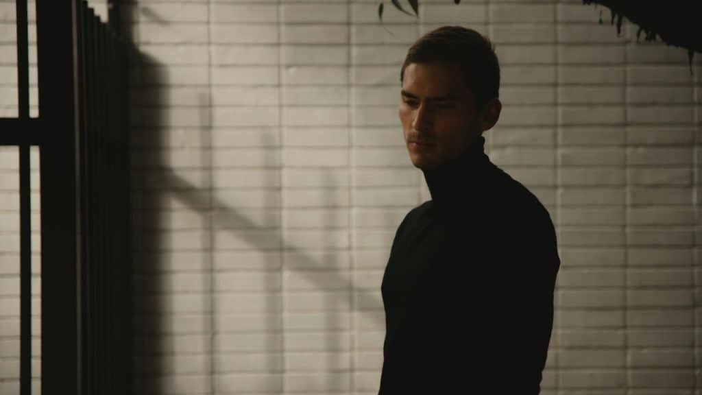 A man in a black turtleneck stands outside against a white brick wall in soft light.