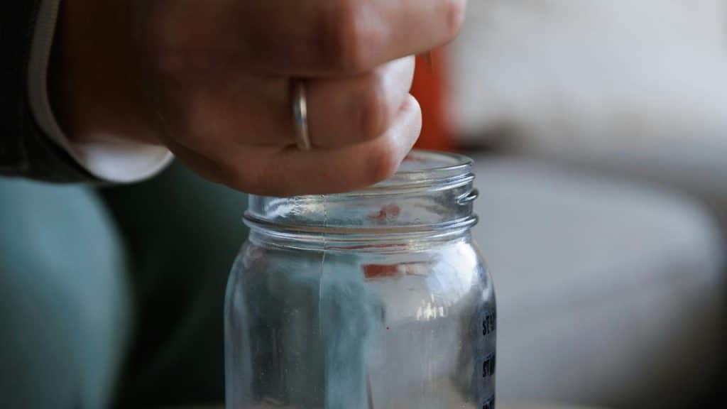 A person placing money into a glass jar savings container.