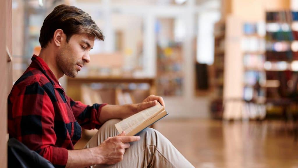 A man sitting on the floor of a library reading a book.
