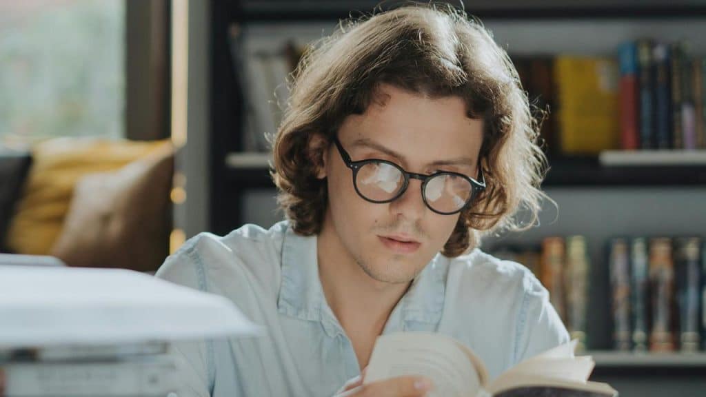 A person wearing glasses reading a book in front of a bookshelf.