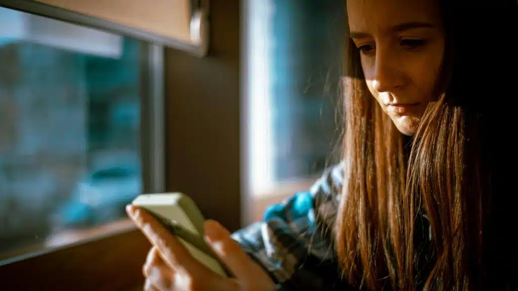A woman looking down at her smartphone by a softly lit window.
