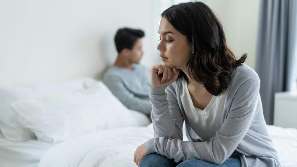 A woman looking upset while sitting on the edge of a bed as her husband lies on the bed in the background.