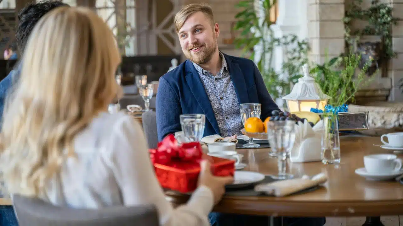 A man smiling at a woman holding a red gift box at a restaurant table.