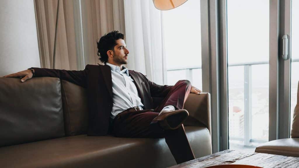 A man in a suit lounges on a sofa by large windows.