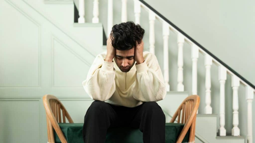 Young man sitting on a chair with his head in his hands, looking distressed, in front of stairs.
