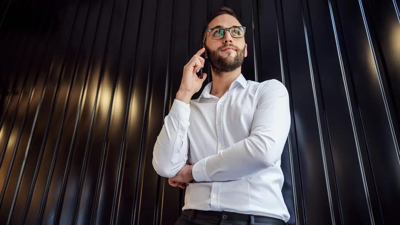 A man in a white shirt and glasses talking on his phone in front of a dark wall.