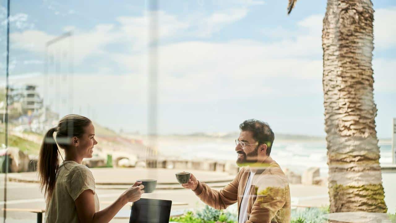 A man and woman smiling and having coffee at a beachside café.