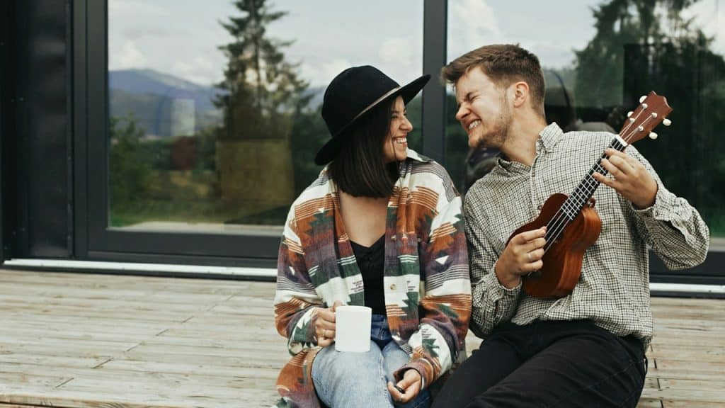 A man playing a ukulele for his stylish woman relaxing on the wooden porch of a modern cabin with big windows in the mountains.