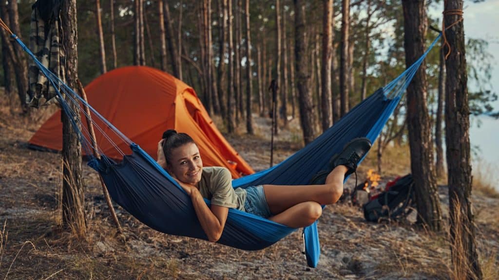 A young woman with her hair in a bun is smiling while relaxing in a blue hammock at a campsite with an orange tent in the background.
