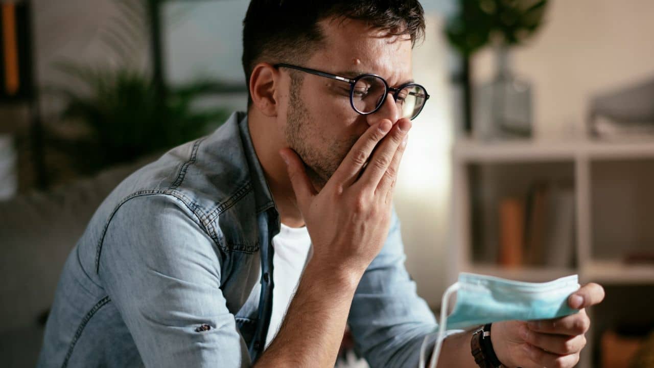Man looking worried, holding a face mask and covering his mouth with his hand.