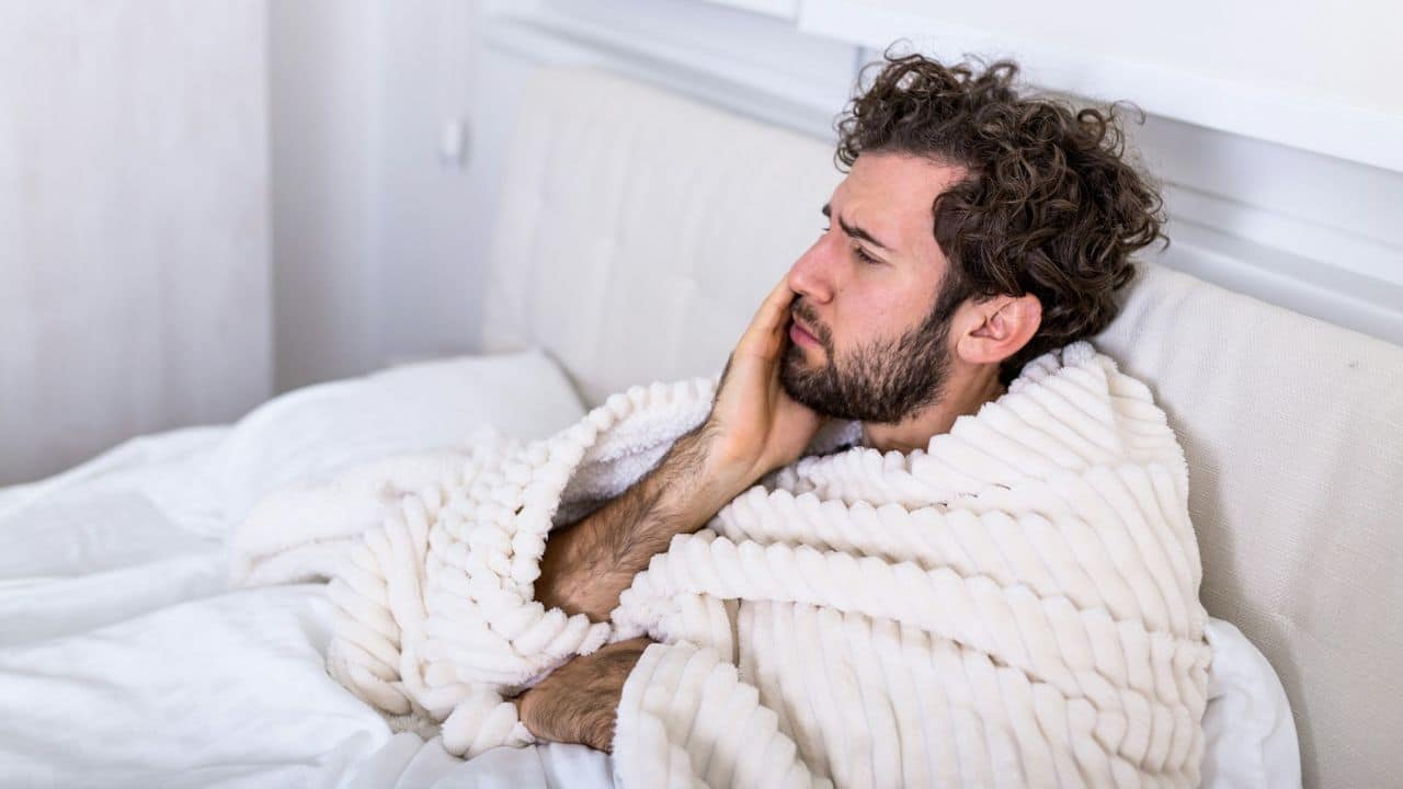 Man with curly hair lying in bed, covered in a white fluffy blanket, looking pensive.