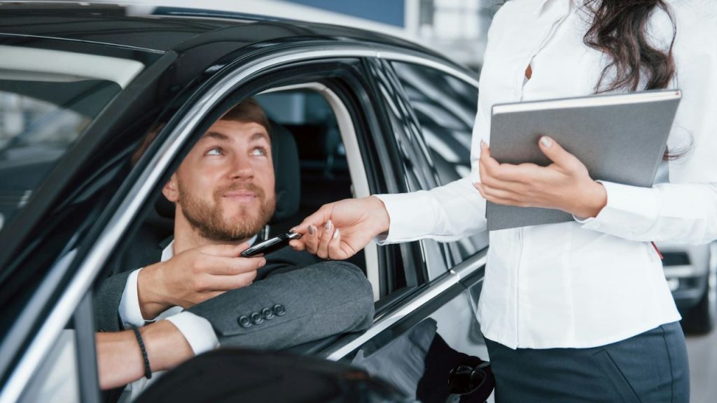 Man in a suit sits in a new car and receives keys from a woman holding a notebook.