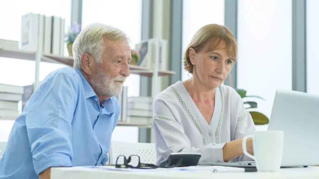 An older couple is intently looking at a laptop computer while sitting at a white desk.