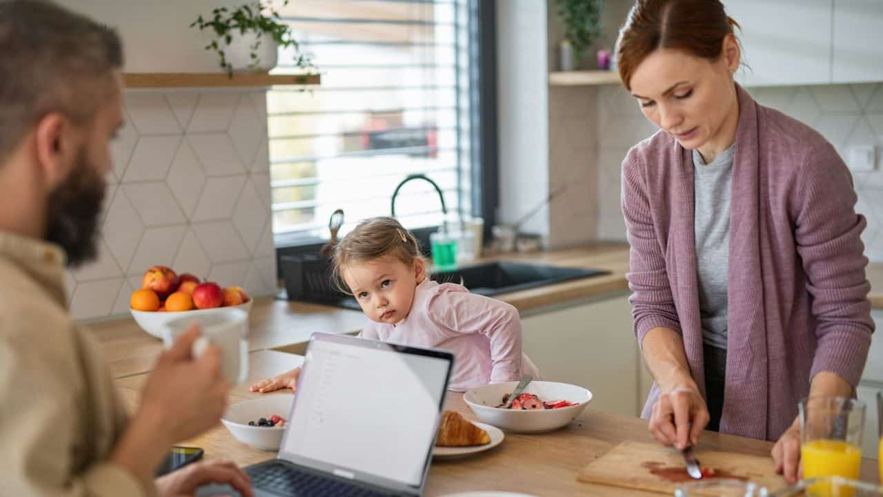 A mother cuts food in a kitchen while a father works on a laptop and a child sits nearby.
