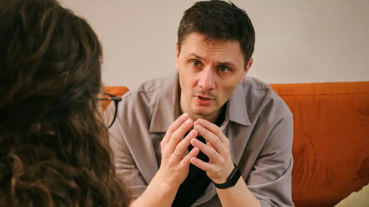 A man in a beige shirt sits, gesturing with his hands, facing an unseen person.