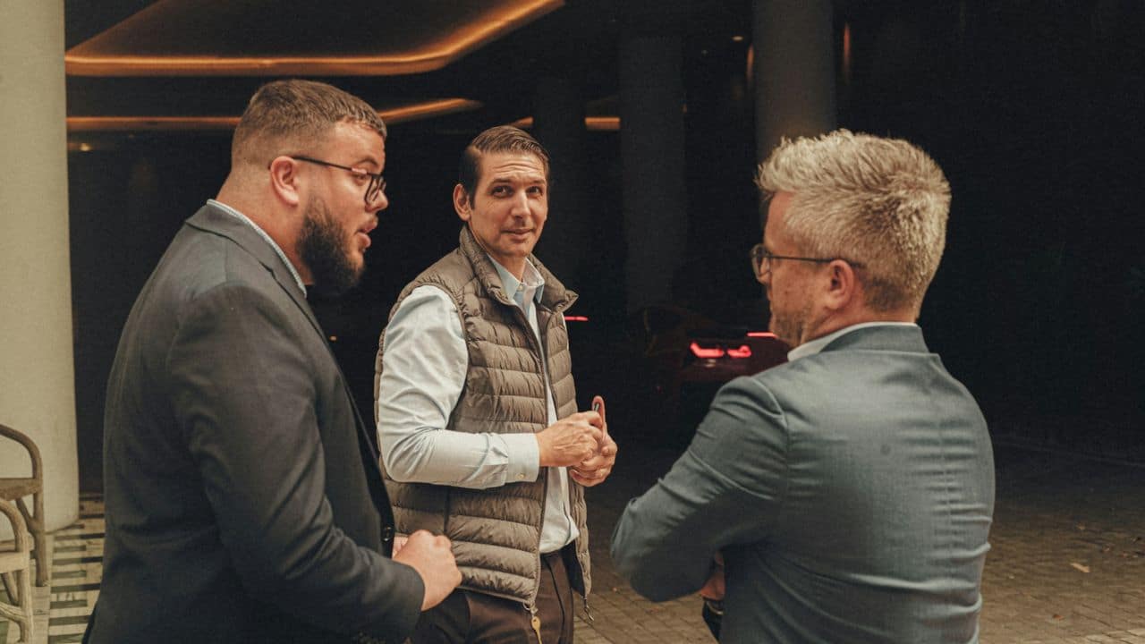 Three men in business attire talking outside at night under a building awning.