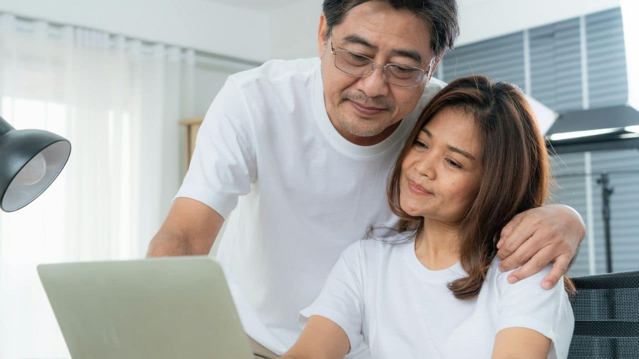 Asian couple in white shirts looking at a laptop together, with the man's arm around the woman.