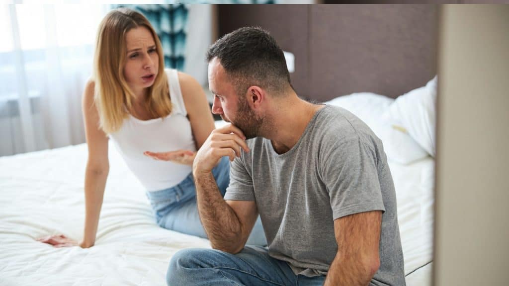 A displeased blonde lady is sitting on a bed and talking with her husband while the man is touching his chin.