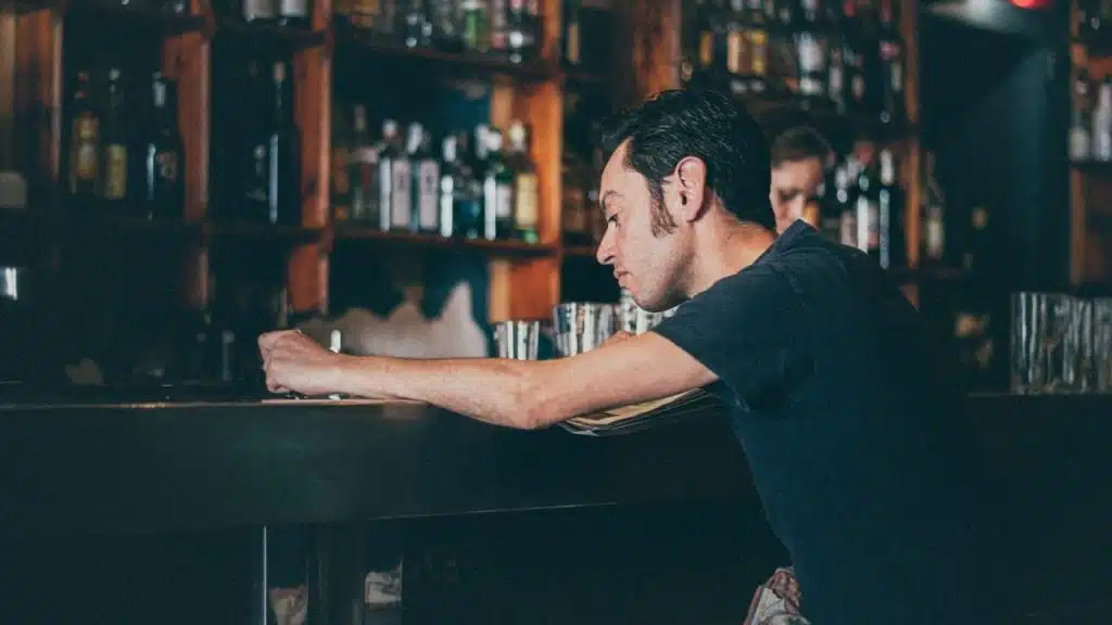 A man in a dark shirt leans on a dark wooden bar in front of shelves of bottles.