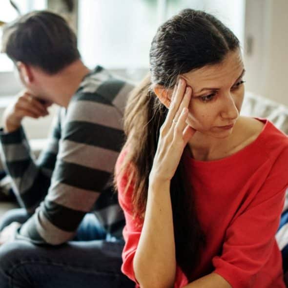 A distressed woman in a red shirt sits with her hand on her forehead, a man facing away in the background.