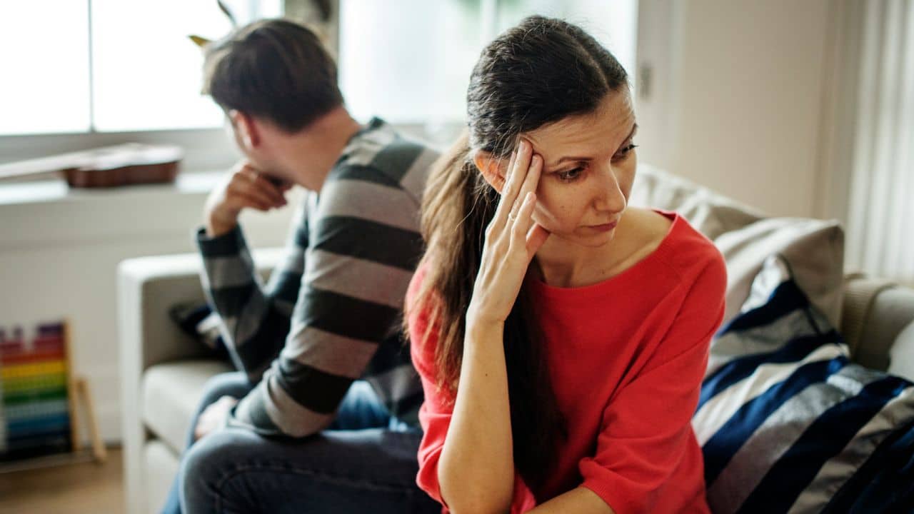 A distressed woman in a red shirt sits with her hand on her forehead, a man facing away in the background.