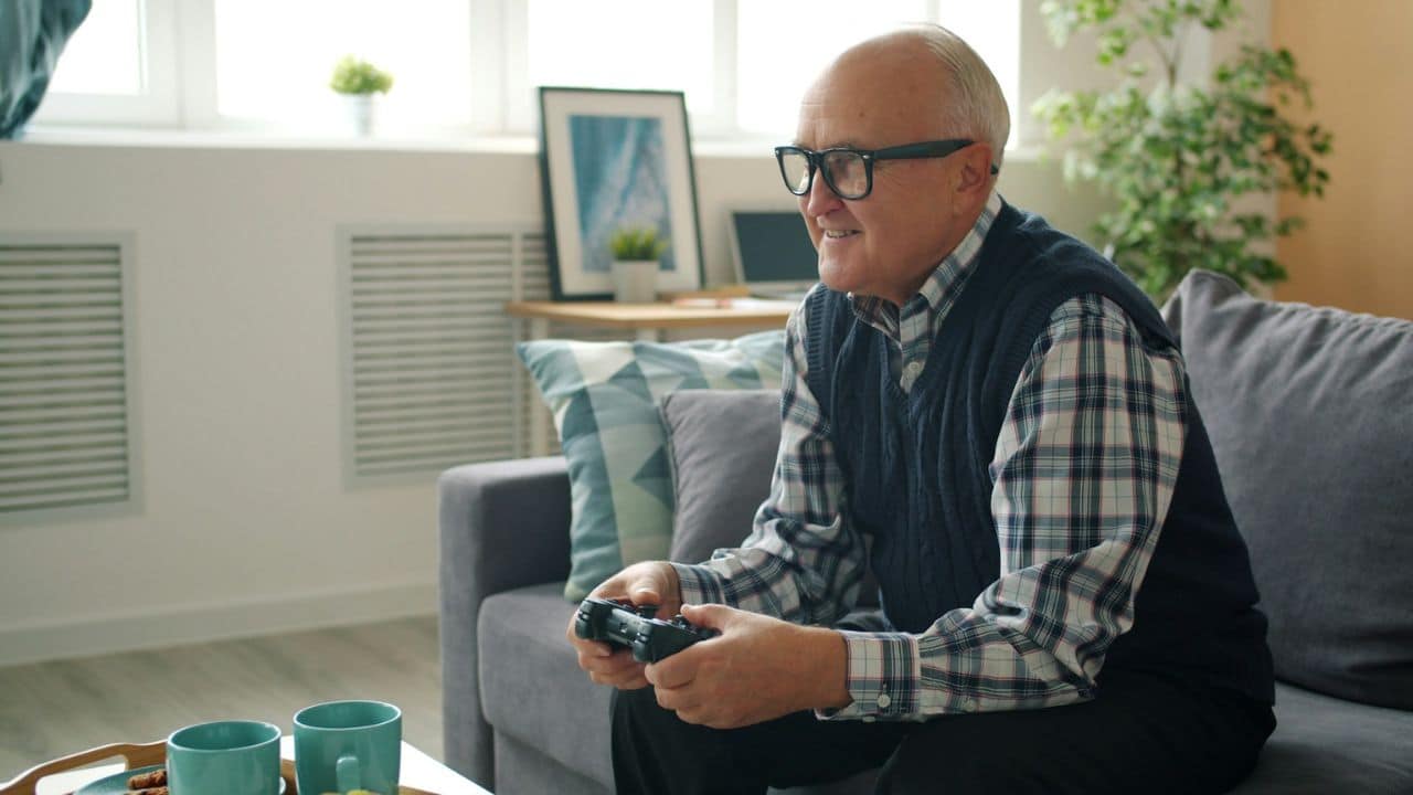An older man smiles while playing a video game console on a couch.